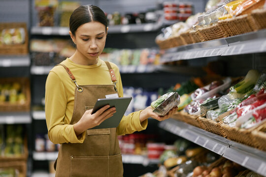 Woman in apricot apron holding tablet while inspecting fresh vegetables in grocery aisle of supermarket. Focused on quality of produce while surrounded by diverse food options - Powered by Adobe