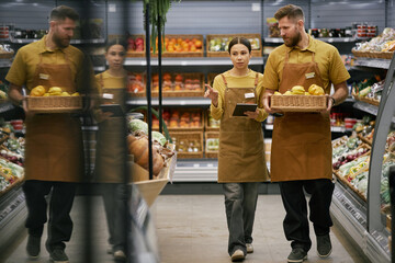 Two grocery store workers are carrying a basket and clipboard while discussing inventory in aisles filled with various fresh produce and goods