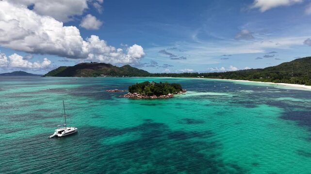 Praslin Island, Seychelles, featuring a tiny rocky islet with palm trees surrounded by crystal-clear turquoise ocean and a nearby sailboat.
