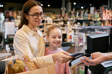 Portrait of woman with her child smiling while paying with smartphone at grocery store checkout...