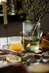 Glass teapot filled with herbal tea containing currant leaves and a golden spoon. A jar of honey and half a lemon are nearby. In the background, a tall candle and a vase with budding branches