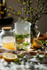 A steaming glass teapot sits on a table, surrounded by fresh lemon halves and a jar of honey.Currant leaves float in the teapot, adding a herbal touch. A lit candle stands nearby, casting a warm glow.