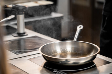 A stainless steel frying pan sits on a stovetop, emitting steam, indicating it is heated and ready for cooking. The kitchen environment is modern and professional.