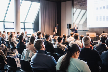 Female speaker giving a talk on corporate business conference. Unrecognizable people in audience at...