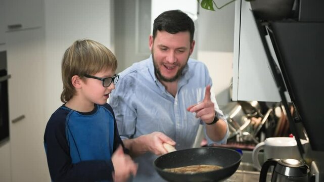 Father flips pancakes in the air, delighting his son on his kitchen. Close-up shot. Slow motion