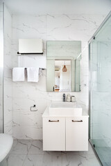 Bright white bathroom with wall-mounted sink, mirror reflecting seaside view, enclosed glass shower, marble tiles, and modern light fixture. Clean, airy, and contemporary space.