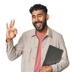 Hispanic male holding laptop in studio cheerful and confident showing ok gesture.