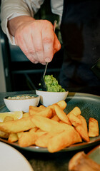A man is preparing food, adding a green sauce to a plate of fries. The plate is on a table, and there are other plates and bowls around it. Scene is casual and relaxed
