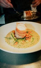 A plate of food with a piece of meat on top of it. The meat is sliced and he is bacon. The plate is set on a table and there is a spoon nearby