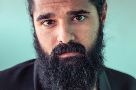Expressive close-up of a man with a full beard and dark eyes holding a contemplative gaze, portraying feelings of introspection and depth, with a clean, soft background contributing to a calm ambiance
