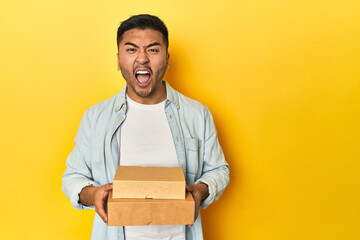 Asian man holding delivery food boxes, yellow studio backdrop screaming very angry and aggressive.