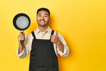 Asian chef holding a pan, yellow studio backdrop smiling and raising thumb up