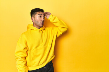 Bold Asian man in yellow hoodie on yellow studio looking far away keeping hand on forehead.