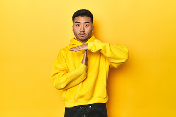 Fototapeta premium Bold Asian man in yellow hoodie on yellow studio showing a timeout gesture.