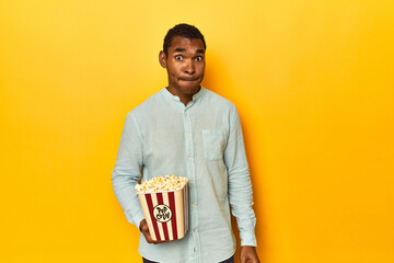 African American man with popcorn, yellow studio, confused, feels doubtful and unsure.