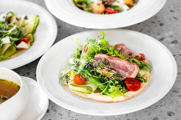 A vibrant salad featuring fresh greens, cherry tomatoes, and slices of ham on a white plate. In the background, other gourmet dishes are displayed, highlighting a beautiful dining setup.