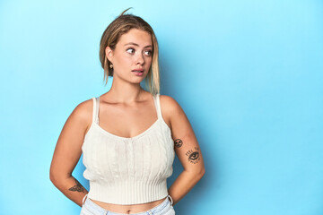 Blonde young woman in white top on blue background being shocked because of something she has seen.