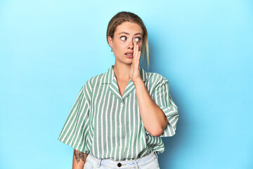 Blonde young woman in green striped shirt on blue background is saying a secret hot braking news...