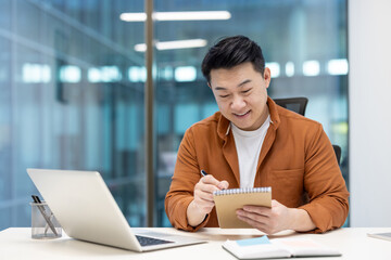 An asian businessman sits at his desk, making notes in a notebook while working on a laptop in a...