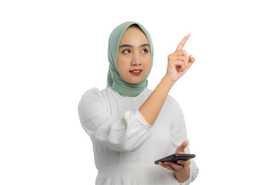 Confident young Asian woman in green hijab and white blouse pointing upward while holding smartphone, showing instruction or idea isolated on transparent background