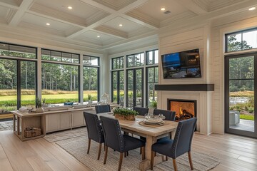 Fototapeta premium Spacious dining and great room with white oak flooring, dark blue chairs, farmhouse-style kitchen chairs, TV above the fireplace, and natural light from large windows overlooking a grassy yard.
