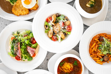 An overhead view of a vibrant assortment of dishes, including a fresh salad, pasta with sauce, soup, and crispy chicken. Each dish is artfully plated on elegant white bowls.