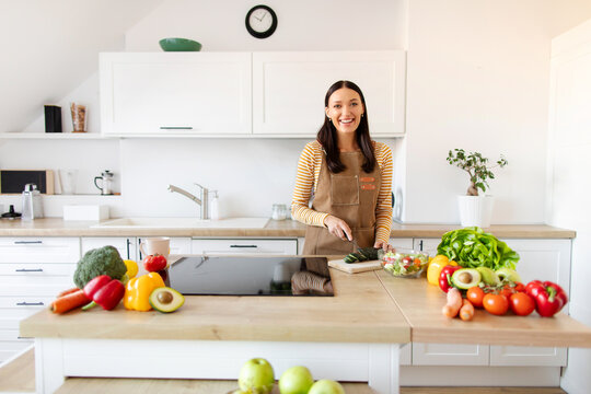 Positive housewife chef lady cutting cucumber for salad, enjoy cooking tasty dinner, woman wearing apron and smiling at camera in modern kitchen