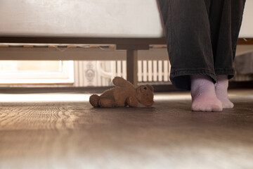 Little girl sitting on bed and under bed forgotten teddy bear at home in bedroom, children's legs
