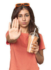 Middle Eastern woman with smoothie standing with outstretched hand showing stop sign, preventing...