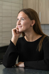 Young woman sits thoughtfully in her kitchen, resting her chin on her hand and gazing upward with a...