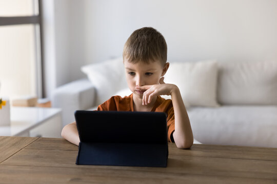 Interactive learning. Focused young kid schoolboy child study from home on distance sit at table read electronic book from tablet computer screen watch video lesson online take part in virtual class