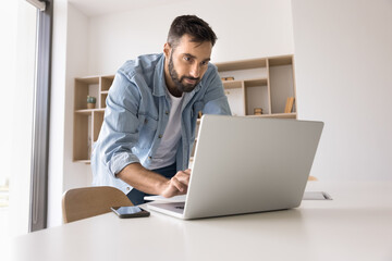 Concentrated young man leaning over work desk with notebook type message in chat write reply to...