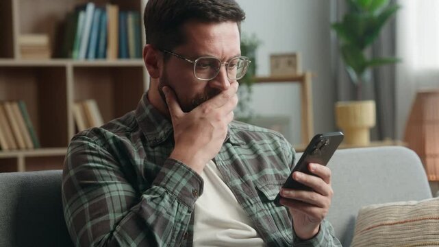 Caucasian adult man male guy at home living room nervous looking at mobile phone bad news amazed shocked close mouth with hand message from bank stressed smartphone notification online error scam