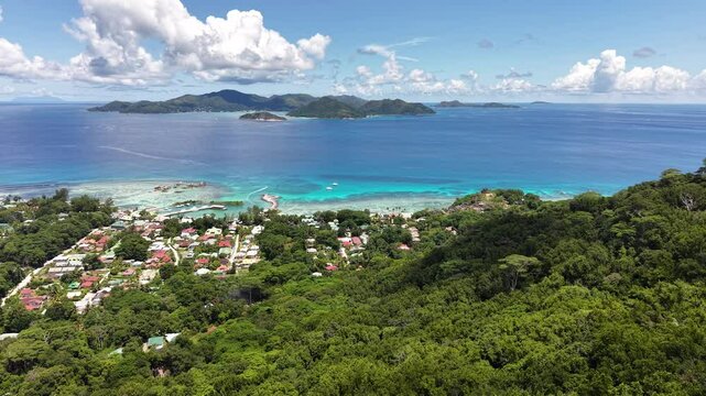 La Digue, Seychelles, viewed from above with a vibrant coastal village, turquoise waters, lush forest, and distant mountainous islands under a bright blue sky.
