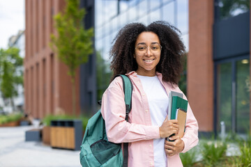 A smiling student stands outside a university building, carrying books and a backpack. Her cheerful...