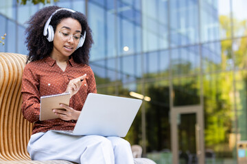 A woman wearing headphones and glasses, takes notes while working on a laptop outdoors. The background has a glass building.
