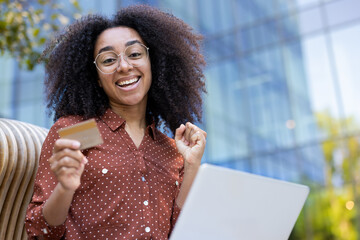 A cheerful woman with curly hair holds a credit card while using a laptop outside, happy about an...