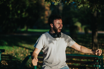 A joyful man with headphones around his neck enthusiastically celebrates in a green park. He...