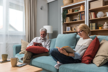 Obraz premium Senior couple sitting at home on sofa, reading a book or online PDF books on digital tablet, checking emails, enjoying simple living and retirement. Mature man and woman love and respect.