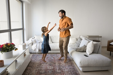 Crazy party. Joyful energetic mature single dad cute preteen kid daughter jumping moving to disco music having fun after doing chores in clean cozy living room interior enjoy weekend at home together