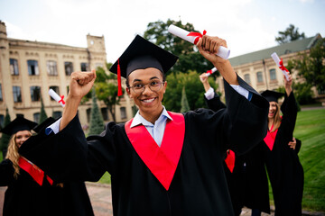 Joyful young graduates celebrating their achievement in academic robes on a sunny day outdoors at a...