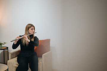 A woman plays the flute while sitting on a chair in a stylish and comfortable interior environment, showcasing the art of music and relaxation in a warm and inviting setting. © qunica.com