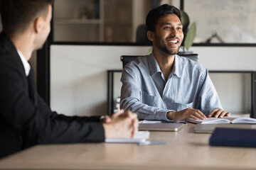 Happy Indian CEO business man holding group meeting in boardroom. Authentic photo of project manager smiling looking at professional team at corporate diverse business team working process