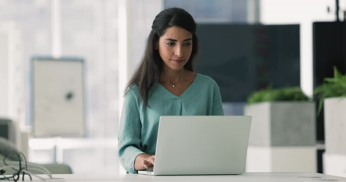 Busy young Latin businesswoman typing on laptop computer in office alone, touching chin, thinking on creative idea for corporate online communication, getting happy, smiling