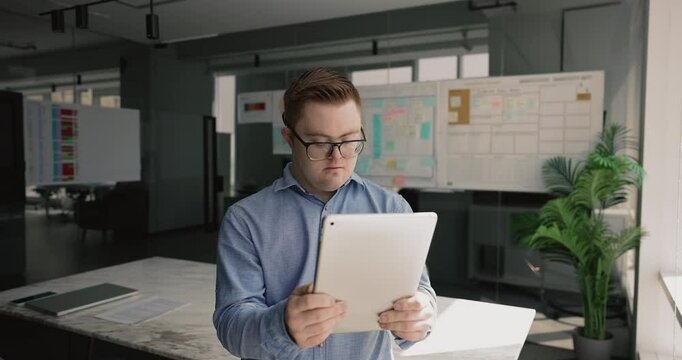 Serious focused down syndrome employee man using tablet in office workspace alone, looking ad gadget, reading or watching online content, thinking on work challenge, problem