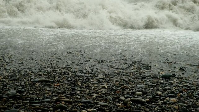 Stormy foamy crushing water waves in sea in overcast weather on pubble beach. Sea storm, power of water and weather, seascape on stone coast. Waves washing stones on coastline.
