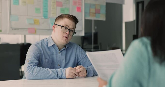 Young down syndrome job candidate man in glasses talking to HR professional woman on interview meeting. Diverse business partners discussing paper contract, agreement, deal