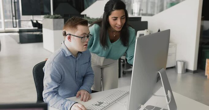 Positive friendly young Latin business woman mentoring down syndrome intern man, helping with corporate work application, software. Diverse colleagues using desktop computer together