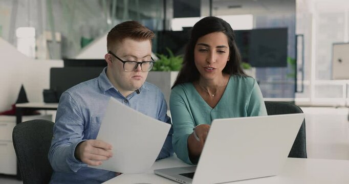 Positive Hispanic project manager woman and down syndrome professional man working at laptop together, checking electronic and document data, analyzing online and paper reports