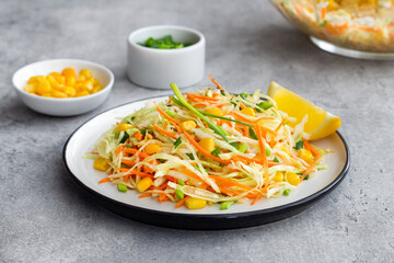 Fresh healthy cabbage salad with carrot, corn and scallions garnished with lemon on white plate on a gray stone background
